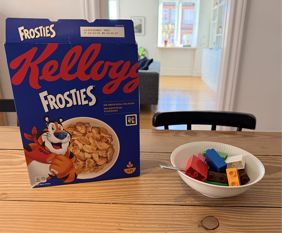 En æske med cornflakes står på en spisebord ved siden af en skål med farverige legoer i.