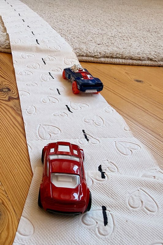 Små legetøjs racerbiler kører på en vej på gulvet lavet af toiletpapir med fartstriber i midten.