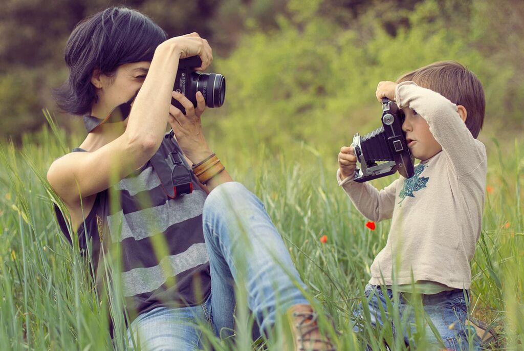 En voksen og et barn fotograferer hinanden i en grøn eng.